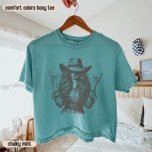 Peut inclure: Un t-shirt boxy bleu sarcelle avec une illustration graphique sombre repr&eacute;sentant une personne aux longs cheveux portant un chapeau de cow-boy et faisant des gestes de la main rock and roll. Le t-shirt a un col rond et des manches courtes.