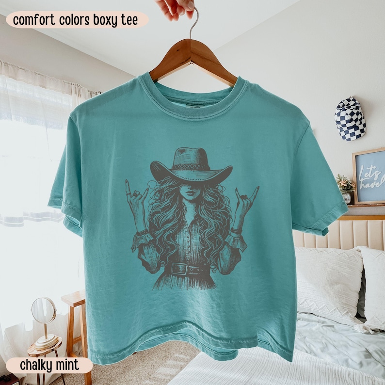 Peut inclure: Un t-shirt boxy bleu sarcelle avec une illustration graphique sombre repr&eacute;sentant une personne aux longs cheveux portant un chapeau de cow-boy et faisant des gestes de la main rock and roll. Le t-shirt a un col rond et des manches courtes.