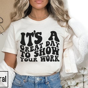 May include: A white t-shirt with black text that reads "It's a great day to show your work" in a retro font.