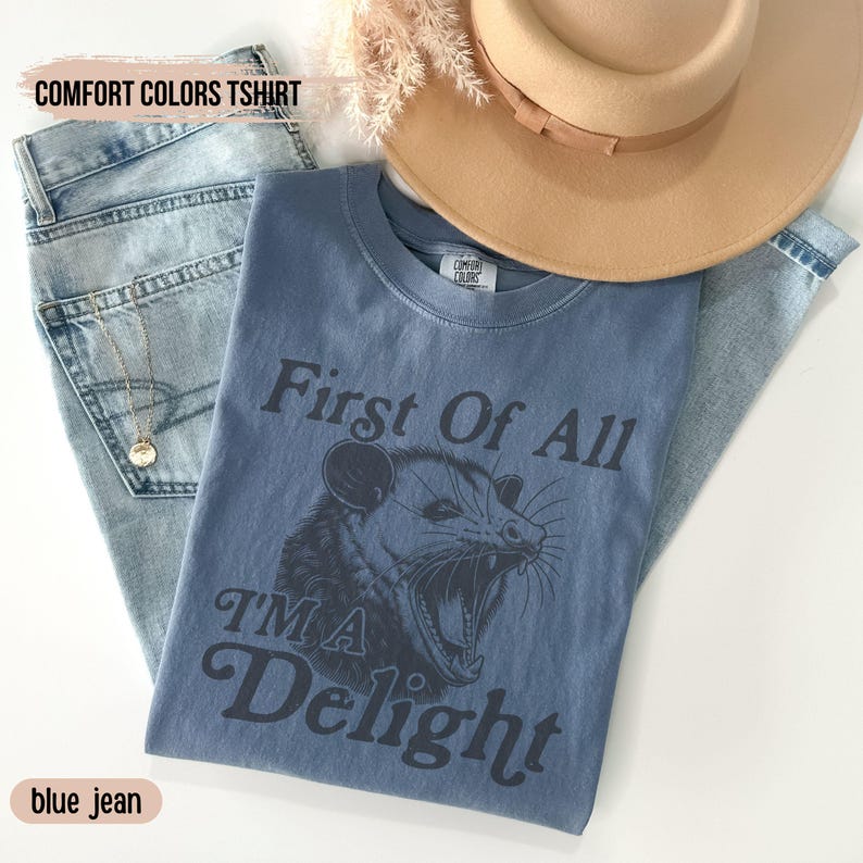 May include: A blue t-shirt with the text "First Of All I'm A Delight" and a black and white illustration of a possum with its mouth open. The shirt is on a blue jean background.