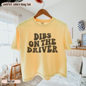K&ouml;nnte beinhalten: Hellgelbes Boxy-T-Shirt mit dem Text "Dibs on the Driver" in Braun, an einem Holzb&uuml;gel h&auml;ngend. Das Shirt geh&ouml;rt zur Comfort Colors Kollektion.
