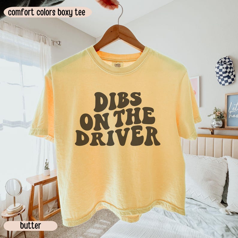 K&ouml;nnte beinhalten: Hellgelbes Boxy-T-Shirt mit dem Text "Dibs on the Driver" in Braun, an einem Holzb&uuml;gel h&auml;ngend. Das Shirt geh&ouml;rt zur Comfort Colors Kollektion.