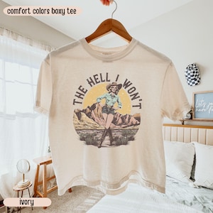 May include: Ivory-colored boxy tee with the text "THE HELL I WON'T" and a graphic of a woman in a cowboy hat. The graphic is set against a desert landscape with a yellow sun. The shirt is hanging on a wooden hanger.