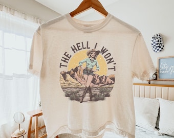 Könnte beinhalten: Elfenbeinfarbenes Boxy-T-Shirt mit dem Text "THE HELL I WON'T" und einer Grafik einer Frau mit Cowboyhut. Die Grafik ist vor einer Wüstenlandschaft mit gelber Sonne platziert. Das Shirt hängt an einem Holzkleiderbügel.