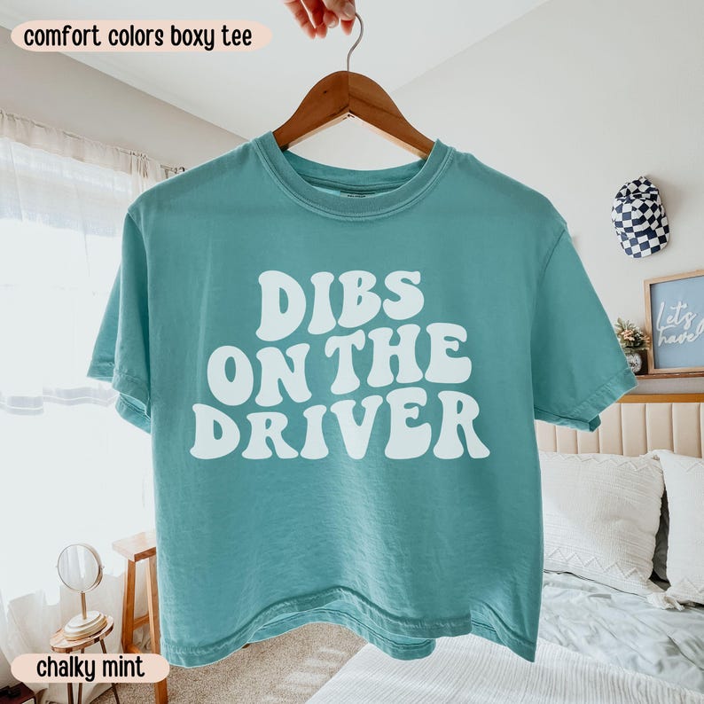 K&ouml;nnte beinhalten: Ein mintfarbenes, kastenf&ouml;rmiges T-Shirt mit dem Schriftzug "DIBS ON THE DRIVER" in wei&szlig;en, fetten, blasenartigen Buchstaben. Das Shirt h&auml;ngt an einem Holzb&uuml;gel. Die W&ouml;rter "comfort colors boxy tee" und "chalky mint" sind im Bild.