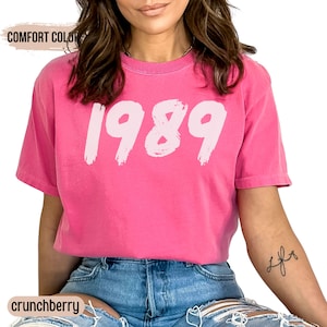 May include: A pink t-shirt with the year 1989 printed in white on the front. The shirt is made of a soft, comfortable fabric and has a relaxed fit.