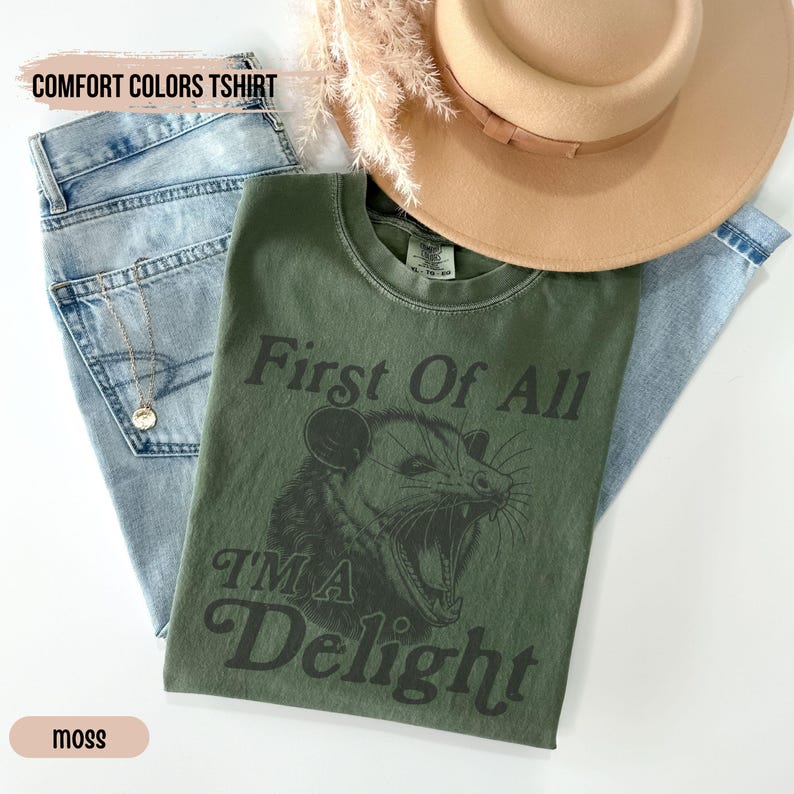 May include: A green t-shirt with the text "First Of All I'm A Delight" and a black and white illustration of an opossum. The shirt is laid on top of blue jeans and a brown hat.