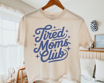 May include: Ivory-colored boxy tee shirt with the text "Tired Moms Club" in blue script. The shirt is hanging on a wooden hanger. The words "comfort colors boxy tee" and "ivory" are also visible.
