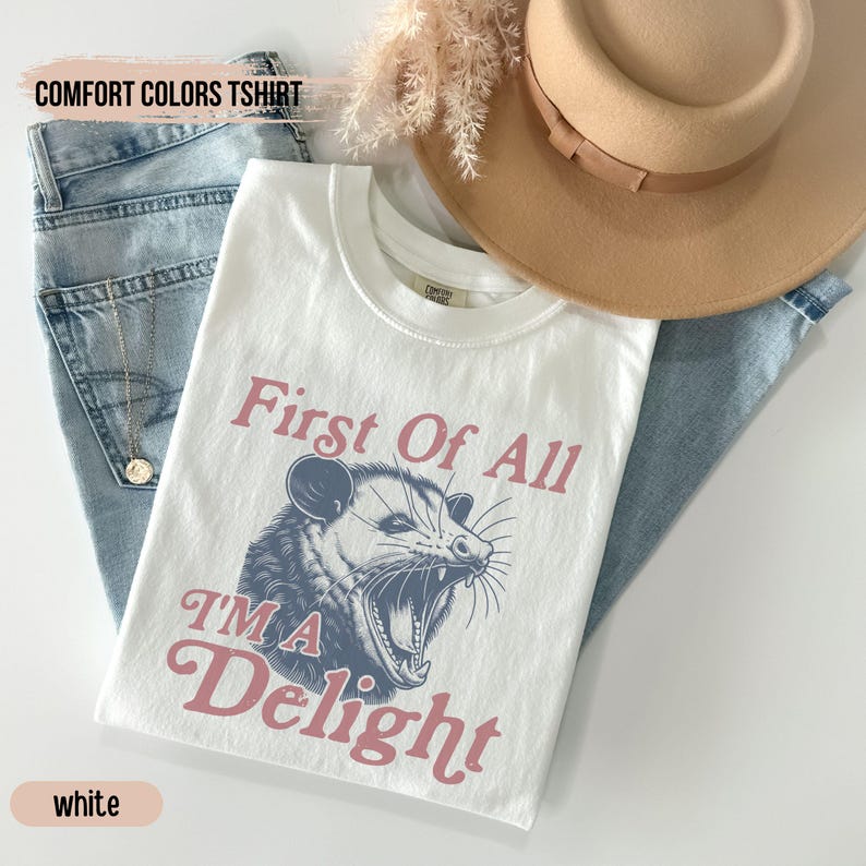 May include: White t-shirt with a graphic of a possum with the text "First Of All I'm A Delight" in pink.