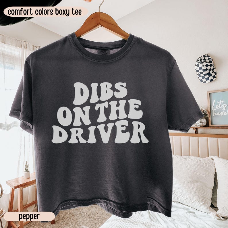K&ouml;nnte beinhalten: Ein dunkelgraues, kastenf&ouml;rmiges T-Shirt mit den Worten "DIBS ON THE DRIVER" in wei&szlig;en, fetten Buchstaben. Das Shirt h&auml;ngt an einem Holzb&uuml;gel. Die Worte "comfort colors boxy tee" stehen oben im Bild, und das Wort "pepper" steht unten.