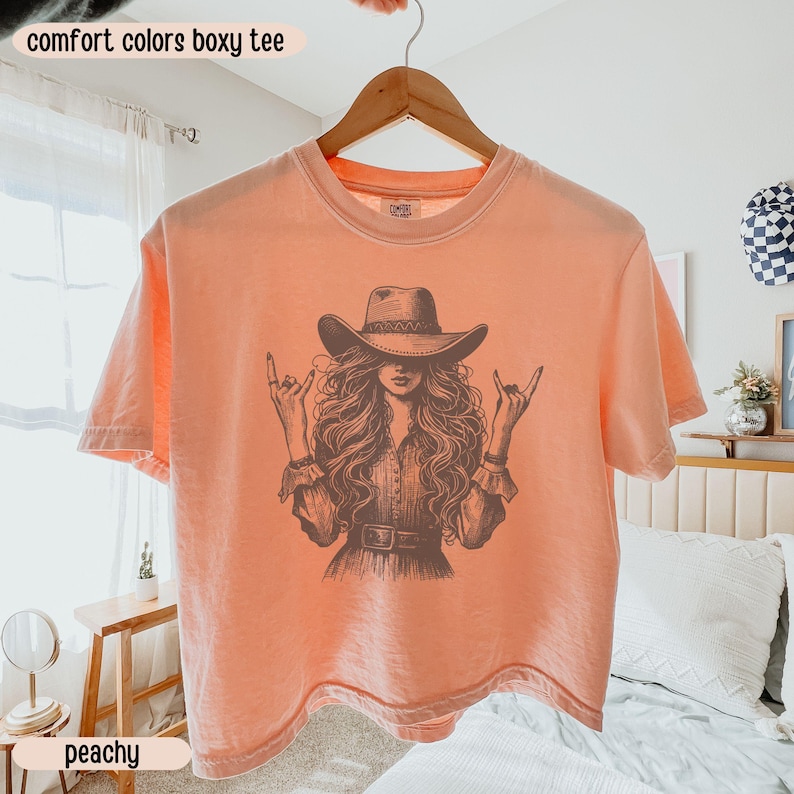 Peut inclure: Un t-shirt boxy couleur p&ecirc;che avec une impression graphique marron fonc&eacute; repr&eacute;sentant une personne aux longs cheveux ondul&eacute;s portant un chapeau de cowboy et faisant des gestes de la main rock and roll. Col rond et manches courtes.