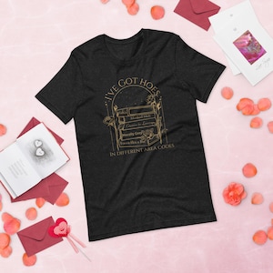 May include: Dark gray t-shirt with a gold graphic that reads "I'VE GOT HOES" above a stack of books with titles like "Masked Men" and "Enemies to Lovers." The text "IN DIFFERENT AREA CODES" is below the books. The shirt is surrounded by pink rose petals and cards.