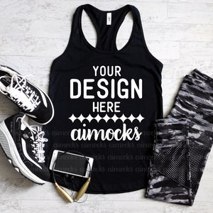 May include: Black racerback tank top with the text "YOUR DESIGN HERE aimocks" in white. The tank top is laying flat on a white wood surface with black and gray leggings, black sneakers, a black cell phone, and gold wireless headphones.