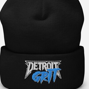 Op de afbeelding: Zwarte beanie met de geborduurde tekst "DETROIT GRIT" in wit en blauw. De hoed is gemaakt van een zacht, gebreid materiaal en heeft een omgeslagen rand. De tekst is in een gestileerd lettertype.