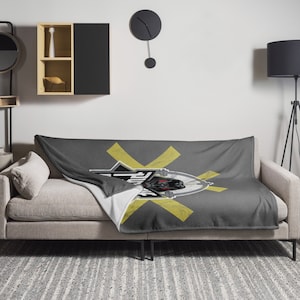 Może przedstawiać: Szary polarowy koc z żółtym X i czarno-białym nadrukiem głowy robota. Koc jest złożony i leży na szarej kanapie.