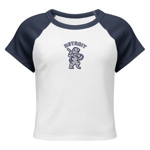 Op de afbeelding: Witte crop top met marineblauwe raglanmouwen. De shirt heeft een Detroit-afbeelding met een honkbalknuppel en het woord "DETROIT" boven de afbeelding. De shirt is gemaakt van een zacht, comfortabel materiaal.