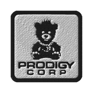 May include: Embroidered patch featuring a black teddy bear design with the text "PRODIGY CORP" in white. The patch has a square shape with rounded corners and a textured, gray background.