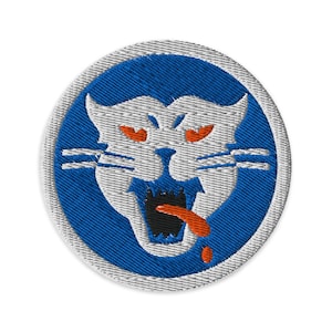 May include: Embroidered patch featuring a stylized white cat face with orange eyes and a red tongue, set against a blue background. The patch has a circular shape with a gray border. The cat's mouth is open, and it has whiskers.