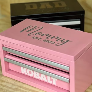 Puede incluir: Caja de herramientas rosa con la palabra "Mommy" y "EST. 2023" impresas en la parte delantera. La caja de herramientas tiene dos cajones con asas plateadas y la palabra "KOBALT" impresa en el cajón inferior. Una caja de herramientas negra con "DAD" y "EST. 2023" está en el fondo.