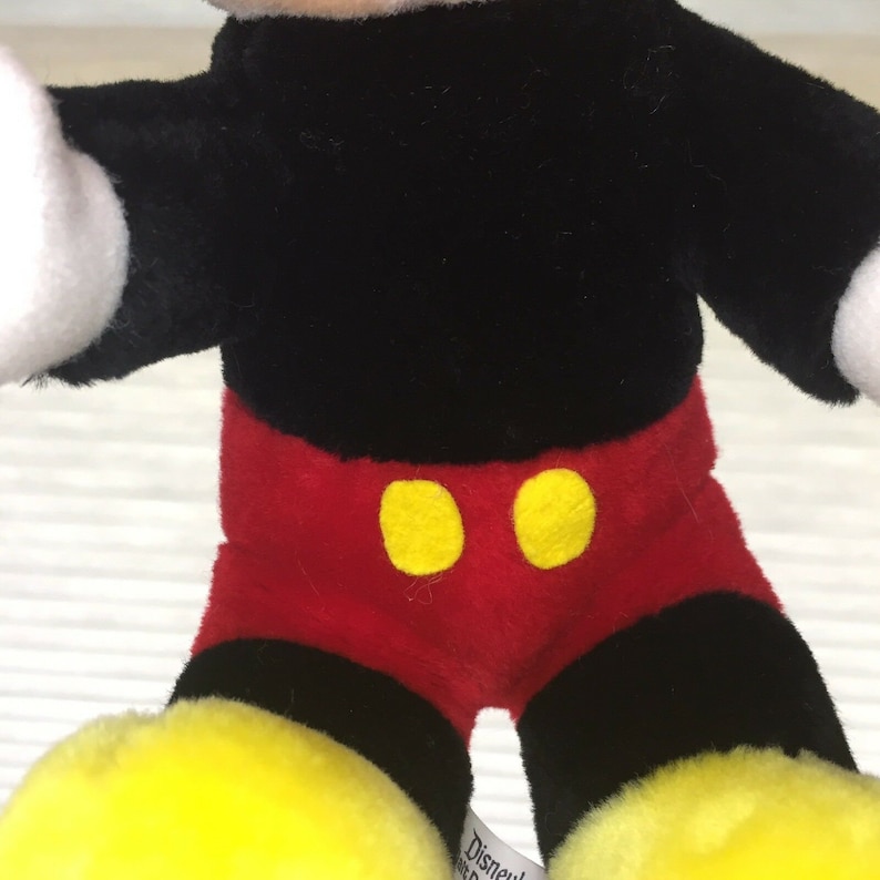 May include: A plush Mickey Mouse toy with black fur, red shorts, and yellow buttons. The toy is sitting with its legs crossed and has a yellow tag on its foot that says "Disney".