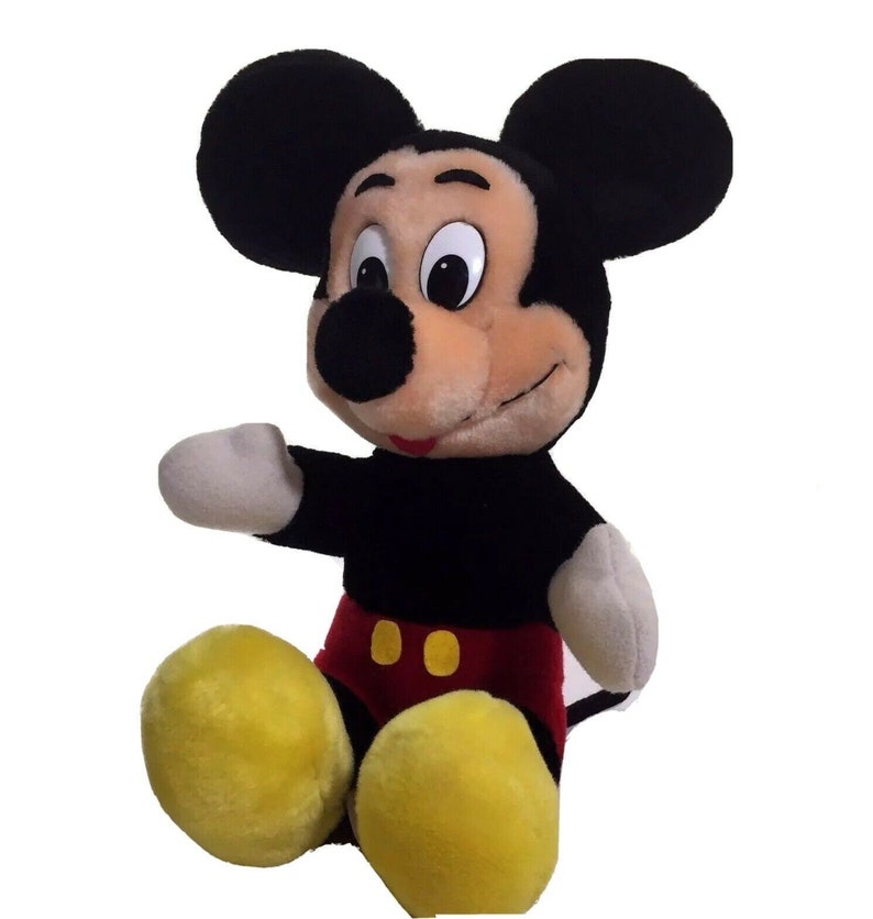 May include: A plush Mickey Mouse toy sitting with his legs crossed. He has black ears, a black nose, and a red and yellow outfit.
