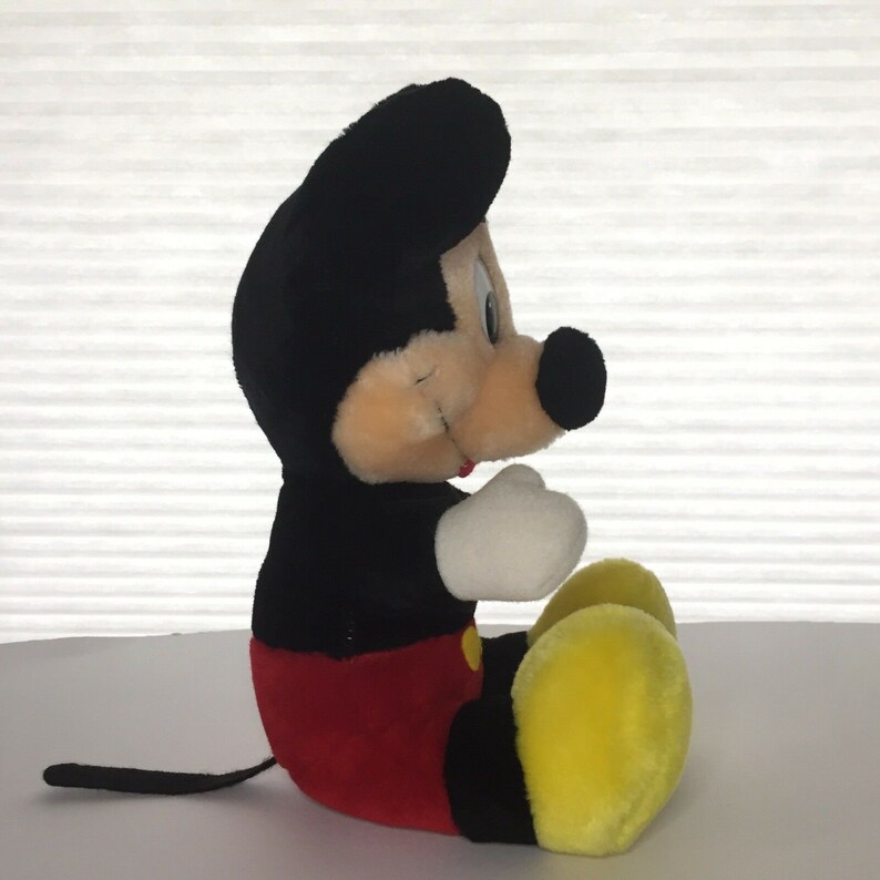 May include: A plush Mickey Mouse toy sitting with his legs crossed. He is wearing red shorts and yellow shoes. His fur is black with a white face and large black ears.