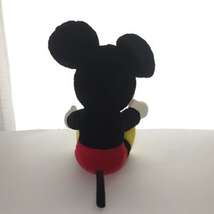 May include: A black plush Mickey Mouse toy sitting with its back to the camera. The toy has red shorts, yellow shoes, and a black tail.