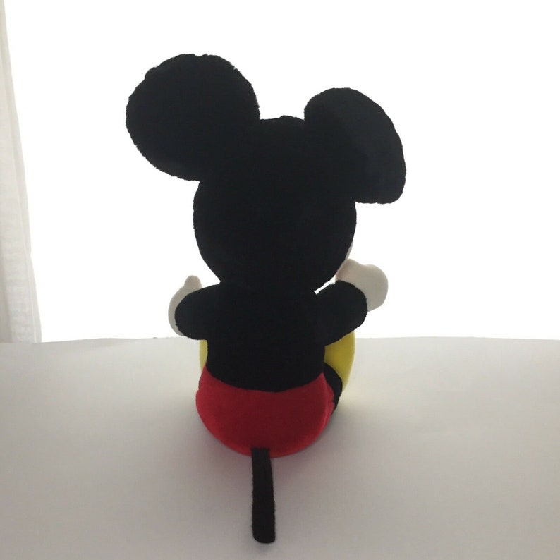 May include: A black plush Mickey Mouse toy sitting with its back to the camera. The toy has red shorts, yellow shoes, and a black tail.