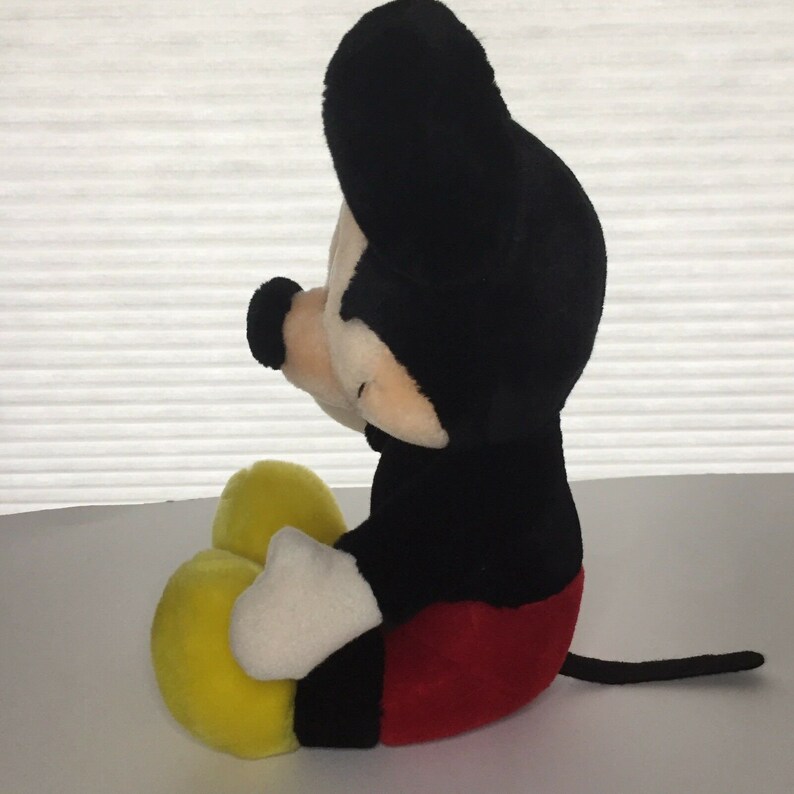 May include: A plush Mickey Mouse toy sitting with his legs crossed. He is wearing red shorts and yellow shoes. His fur is black and his face is white with black eyes and a black nose.