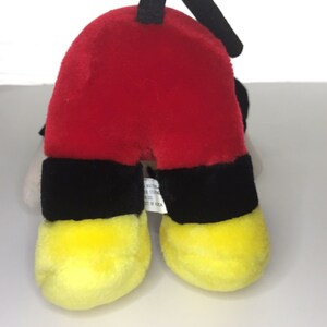 May include: A red, black, and yellow plush toy of Mickey Mouse. The toy is lying on its back with its feet in the air. The toy has a black tag on its back that says "Material: Polyester, Stuffing: Polyester Fiber, Made in Korea."