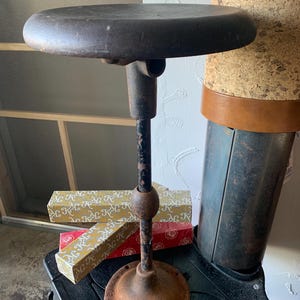 May include: A vintage industrial stool with a round wooden seat and a black metal base. The base has a ball joint and a wide, rusty foot.