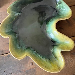 May include: A leaf-shaped ceramic serving dish with a handle. The dish features a glossy glaze in shades of green and yellow, with a dark green center and lighter edges. The dish is placed on a wooden surface.