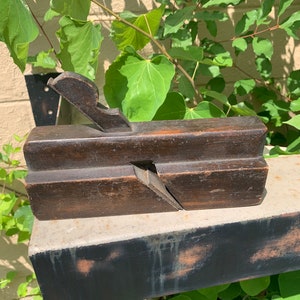 May include: A vintage wooden hand plane with a dark brown finish. The plane has a wooden handle and a metal blade. The plane is sitting on a wooden surface.