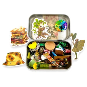 Puede incluir: Una colección de objetos caprichosos, que incluyen pegatinas, amuletos y pequeñas baratijas, se exhibe alrededor de una caja de metal abierta. Los artículos incluyen una pila de libros, un sombrero de girasol, un personaje parecido a un árbol, un hada y un diseño de árbol arcoíris.