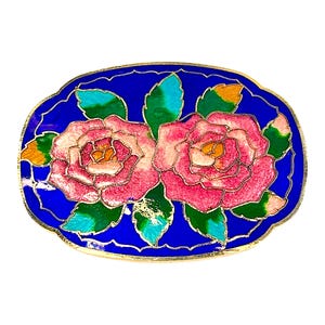 Puede incluir: Broche ovalado de cloisonné con dos rosas rosas con hojas verdes sobre un fondo azul vibrante. Las rosas tienen centros amarillos y están delineadas en dorado. El broche tiene un borde dorado.