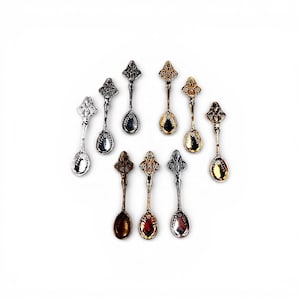 May include: An assortment of decorative spoons in various metallic finishes, including silver, gold, and bronze. Each spoon features an ornate, detailed handle and a small, oval bowl. The spoons are arranged in a circular pattern against a white background.