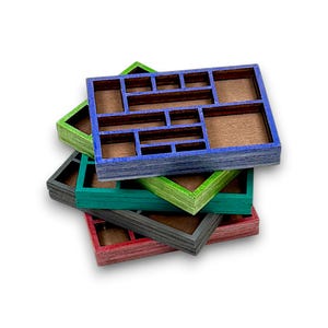 Op de afbeelding: Een stapel van vier rechthoekige houten organisertrays met meerdere binnenvakken. Elke tray heeft een apart gekleurd frame in blauw, groen, donkergrijs en rood, met een natuurlijke bruine houten bodem zichtbaar in elk compartiment.