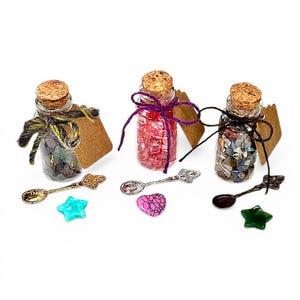 May include: Three small glass bottles with cork stoppers, filled with colorful stones. Each bottle is tied with twine and has a blank tag. Decorative spoons and star and heart-shaped stones are arranged in front.