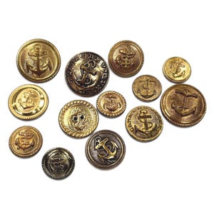 May include: A collection of twelve round gold-toned metal buttons of varying sizes, each featuring a raised anchor design. Some buttons have a rope-like border, while others display embossed text around the central anchor motif.