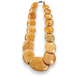 May include: A necklace featuring large, oval-shaped beads in varying shades of tan and cream. The beads are strung together with a dark metal clasp and a small wooden bead. The necklace has a natural, earthy aesthetic.