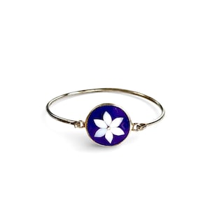 May include: Silver-tone bangle bracelet with a circular charm. The charm features a white flower design set against a deep blue background. The bracelet is a simple, elegant piece of jewelry.