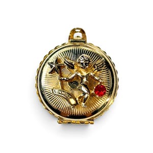 May include: A round gold-tone metal locket featuring a raised cherub figure holding a star and a red gemstone. The background has a sunburst pattern, and a banner at the bottom is engraved with the word JULY. The locket has a top bail for hanging.