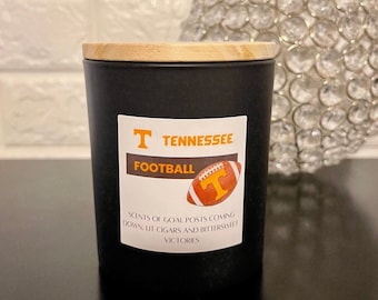 May include: A black candle with a wooden lid. The label reads "Tennessee Football" with a football graphic. The text below says "Scents of goal posts coming down, lit cigars and bittersweet victories."