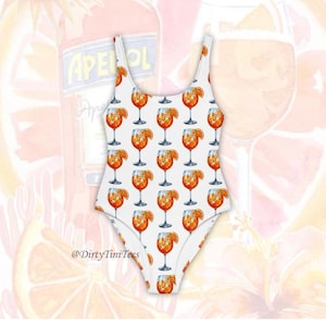 May include: White swimsuit with a repeating pattern of Aperol Spritz cocktails, each with an orange slice. The background shows a blurred Aperol bottle and a large cocktail glass. The text "@DirtyTiniTees" is visible.