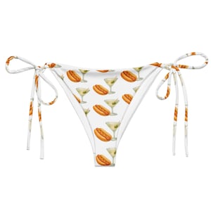 May include: A white bikini bottom featuring a repeating pattern of hot dogs and martini glasses with olives. The garment has thin side ties for adjustment, a low-rise cut, and a clean white background that highlights the colorful food and drink print.