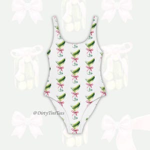 May include: White swimsuit with a pattern of green pickles in martini glasses with pink ribbons. The swimsuit has a scoop neck and a high leg cut. The text "@DirtyTiniTees" is printed on the bottom of the swimsuit.