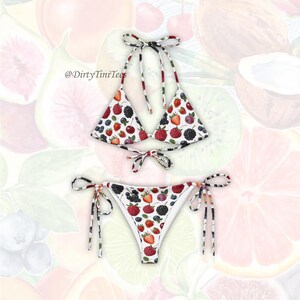 Puede incluir: Conjunto de bikini blanco con estampado de frutas. La parte superior es de estilo halter con cierres de lazo, y la parte inferior es un bikini de cuerda clásico. El estampado presenta fresas, frambuesas, arándanos y moras. El texto "@DirtyTiniTees" es visible.