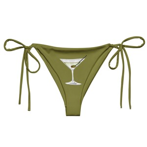 May include: An olive green bikini bottom featuring side tie closures and a centered graphic illustration of a clear martini glass containing a single green olive on a toothpick, rendered in shades of white, grey, and light green.