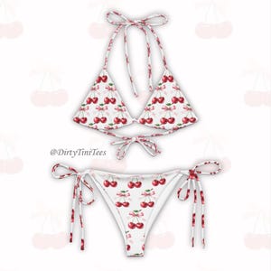 May include: White bikini with a cherry print. The top is a triangle style with adjustable straps. The bottom is a cheeky style with ties on the sides. The bikini is made of a soft, stretchy fabric.