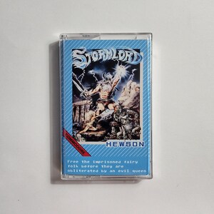 May include: A vintage Commodore game cartridge for the game Stormlord by Hewson. The cartridge is blue and white with a picture of a warrior holding a sword and a lightning bolt. The text on the cartridge reads "Stormlord", "Commodore", and "Hewson". The text below the image reads "Free the imprisoned fairy folk before they are obliterated by an evil queen."